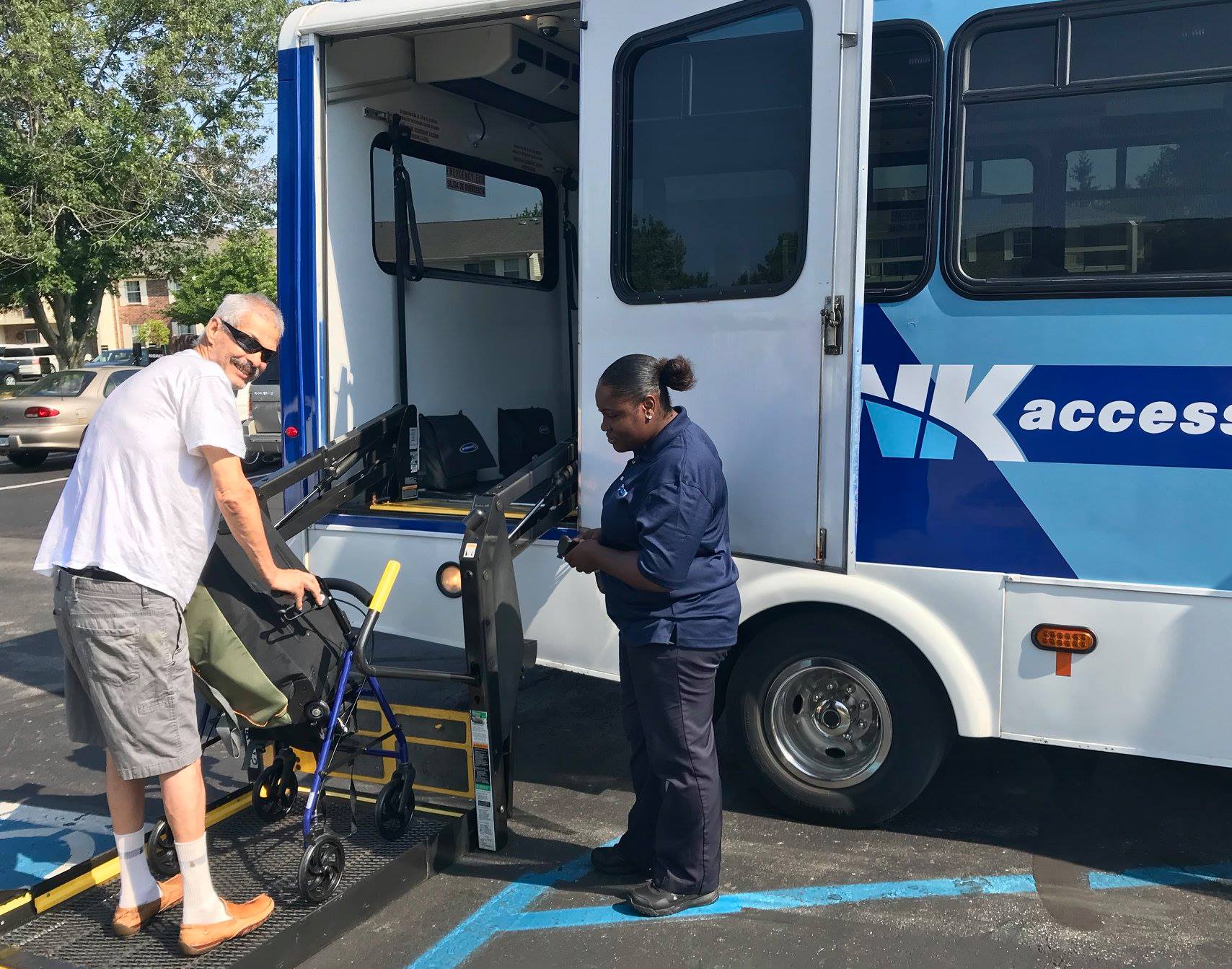 A man with a walker stands on a lift to board the bus while the driver operates the lift controls